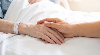 Primer plano de dos manos, una con pulsera blanca, entrelazadas sobre una sábana blanca. Al fondo, un pijama azul de hospital y una cama blanca.