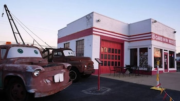 Dos grúas oxidadas con ojos grandes y dientes, una con grúa, estacionadas frente a un edificio blanco y rojo con un letrero 'Cars on the Route 66' y mesas