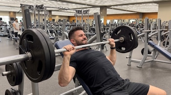 Un hombre de 30 años realiza press de banca inclinado con barra y pesas en un gimnasio. Se observan otras máquinas de ejercicio y personas en el fondo.