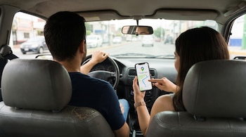 Vista interior de un vehículo con un hombre al volante y una mujer sujetando un teléfono inteligente que muestra la aplicación Google Maps.