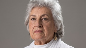 Primer plano del rostro de Flor Vargas, una mujer mayor de cabello gris rizado, con arrugas visibles, labios rosados y pendientes dorados en forma de lágrima, sobre fondo gris.