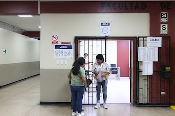 Dos mujeres y un hombre en el umbral de un aula identificada como "AULA 401" en la UNMSM, con carteles electorales y de seguridad visibles