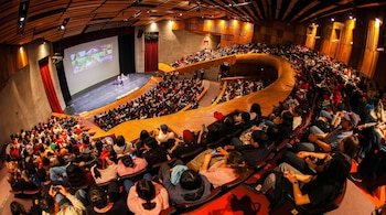 Auditorio lleno de personas sentadas, escenario con dos personas y una mesa, gran pantalla proyectando nueve recuadros con rostros.