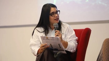 Una mujer con anteojos y camisa blanca habla por un micrófono mientras sostiene un informe impreso sentada en un sillón rojo