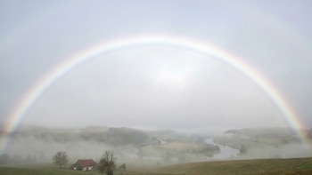 Arcoíris sobre paisaje con rocas, vegetación, río sinuoso, un pueblo con casas de colores y columnas de vapor