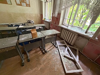 Interior de un aula de orfanato con pupitres cubiertos de cristales rotos, ventanas destrozadas y un marco de ventana caído en el suelo