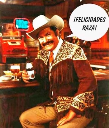 Un hombre con sombrero vaquero y chaqueta bordada sostiene una botella en un bar con un bocadillo de diálogo que dice ¡Felicidades raza!
