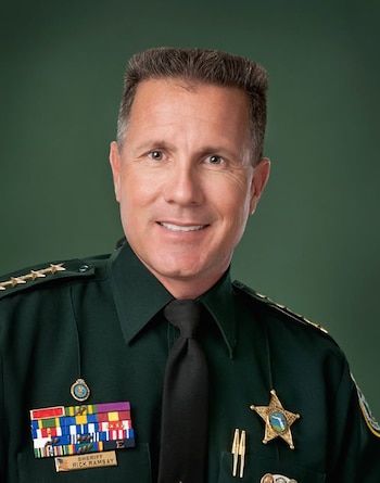 Retrato de Rick Ramsay con uniforme verde de sheriff, insignias, condecoraciones y una placa dorada sobre un fondo verde