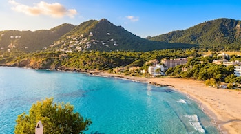 Playa de arena con bañistas, mar turquesa, hoteles y casas en la costa y en montañas cubiertas de pinos. Cielo azul y algunas nubes.