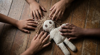Seis brazos de niños rodean una muñeca de trapo beige con ojos de botón y cabello de lana sobre un suelo de madera.