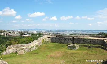 Ruinas de un castillo de piedra con patio de hierba. Al fondo, una ciudad con edificios y árboles, un cuerpo de agua y montañas bajo un cielo azul y nubes
