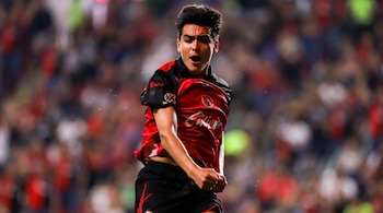 Hombre joven con camiseta roja y negra, pantalones cortos negros, rostro sudoroso y boca abierta en un campo de fútbol con público borroso de fondo.