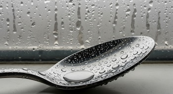 Primer plano de una cuchara de metal brillante cubierta con numerosas gotas de agua, apoyada sobre un alféizar de ventana con un vidrio empañado detrás.