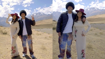 Wendy Sulca con vestido crema y sombrero, y Micro TDH con chaqueta negra y gorro peludo, posan en un paisaje montañoso seco bajo un cielo azul con nubes