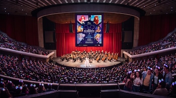 Un auditorio con espectadores usando vinchas de luz, una orquesta sinfónica en el escenario y una pantalla publicitaria de Disney.