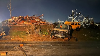 Vista nocturna de un SUV blanco y un Jeep amarillo dañados en una carretera llena de escombros. Al fondo, restos de edificios y árboles rotos