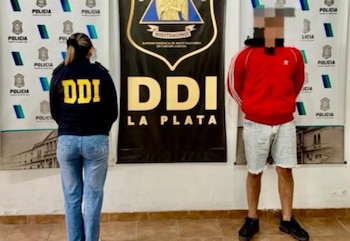 Una mujer con chaqueta azul que dice "DDI" y jeans azules está de espaldas junto a un hombre con sudadera roja y shorts claros. Detrás, carteles de "Policía" y "DDI La Plata"