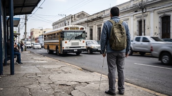Adulto joven de espaldas con mochila y bastón blanco espera en parada de autobús urbano. Autobús y vehículos se mueven en la calle, edificios al fondo. Aceras irregulares.