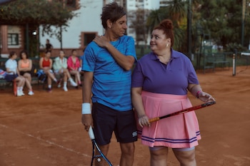 Hombre y mujer en cancha de tenis. Él con camiseta azul y raqueta; ella con camiseta morada, falda rosa y raqueta. Personas sentadas al fondo