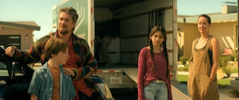 Una mujer (Jena Malone) con overol, un hombre con cárdigan y dos niños posan frente a un camión de mudanzas verde y casas suburbanas