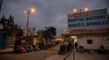 Calle de noche con personas haciendo fila frente al Hospital Nacional Sergio E. Bernales. Farolas encendidas, personas sentadas y acostadas, algunas con mantas.