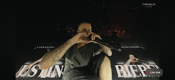 Justin Bieber en el escenario con un micrófono, haciendo un corazón con las manos. Viste ropa oscura, con tatuajes en los brazos y fuegos artificiales al fondo