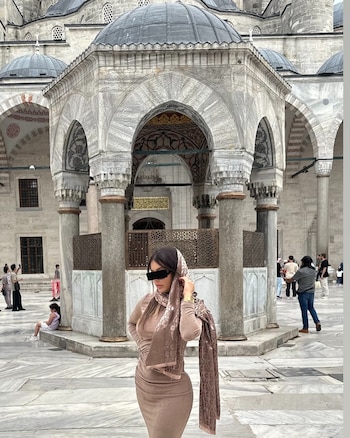 Una mujer con vestido beige, pañuelo en la cabeza y una barra negra sobre los ojos posa en el patio de mármol de una mezquita
