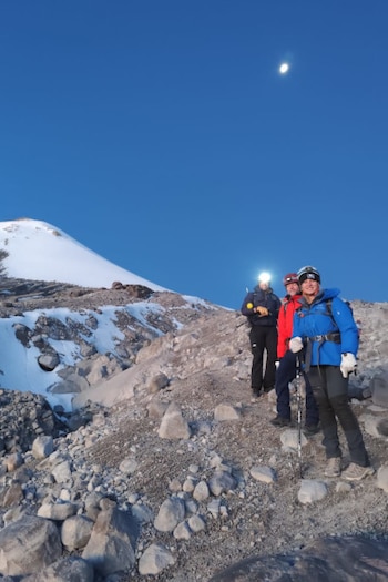 Tres montañistas con ropa de invierno y bastones están en una ladera rocosa con nieve bajo un cielo azul