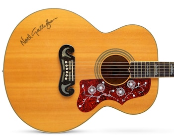 Guitarra acústica de madera clara con la firma de Noel Gallagher, un golpeador rojo decorado con flores blancas y un puente flotante oscuro