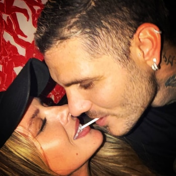 Primer plano de un hombre barbudo y una mujer rubia con gorra, compartiendo un chupetín entre sus labios. El hombre tiene tatuajes en el cuello