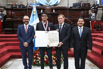 Cuatro hombres con traje posan sonriendo, dos de ellos sostienen un documento de premio. De fondo, una bandera de Guatemala y escaleras rojas