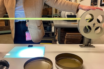Persona examinando una tira de película amarilla en un carrete grande sobre una mesa blanca. Una caja de luz brillante y dos latas de película abiertas están en primer plano