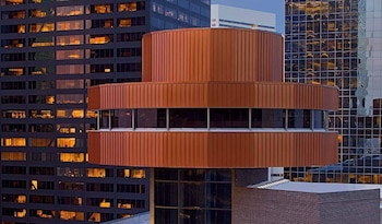Vista cercana de un edificio cilíndrico de color cobre con ventanas horizontales, flanqueado por rascacielos con fachadas de vidrio y luces internas encendidas