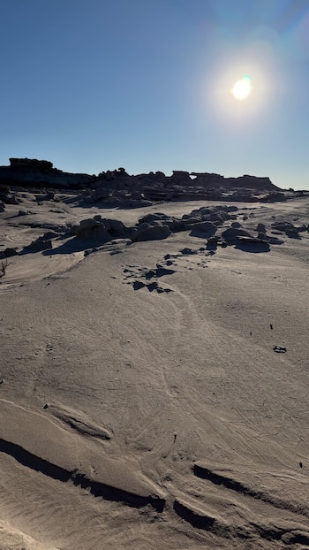 Un cielo azul claro con un sol brillante sobre un paisaje árido de terreno arenoso y formaciones rocosas oscuras que proyectan sombras