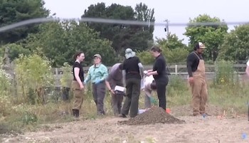 Un grupo de seis personas con ropa de trabajo y guantes, una de ellas con sombrero, examina el suelo removido y una pila de tierra en un terreno abierto con vegetación