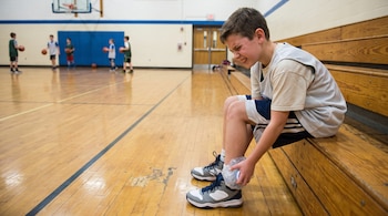 Un niño sentado en un banco de madera de una cancha de baloncesto, con ropa deportiva, sostiene una botella de agua congelada en su tobillo.