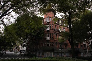 Edificio de ladrillo rojo con torreón y balcones, rodeado de árboles verdes. Calle con vehículos estacionados y algunas personas. Cielo nublado