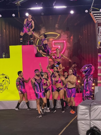 Un grupo de diez atletas en uniformes deportivos morados y rosados posan en un escenario con plataformas de colores, un gran logo "G" y una mascota de dragón