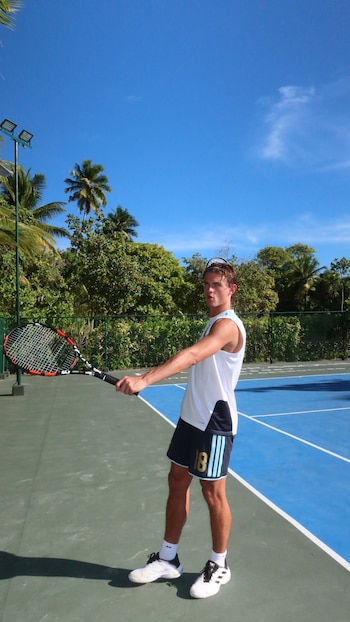Hombre joven en cancha de tenis. Viste camiseta blanca, pantalones cortos azules con el número 18 y zapatillas blancas. Sostiene raqueta. Palmeras y cielo azul