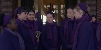Grupo de jóvenes mujeres con abrigos, bufandas y diademas moradas en un interior. Una mujer sostiene un ramo pequeño de flores moradas y blancas
