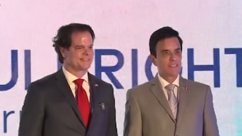 Dos hombres con traje y corbata posan frente a una pantalla con la palabra Fulbright