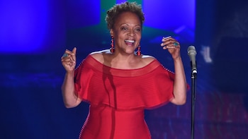 Cassandra Wilson con un vestido rojo sonríe con los brazos levantados junto a un micrófono en un escenario iluminado de azul