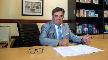 Hombre con traje gris y corbata azul sentado en un escritorio de madera, mirando a la cámara y haciendo un pulgar hacia arriba, con un librero detrás