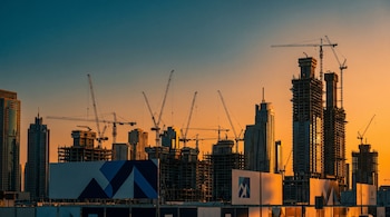 Skyline de una ciudad en construcción al atardecer, mostrando numerosos rascacielos en obra y grúas, con grandes carteles de empresa constructora en primer plano.