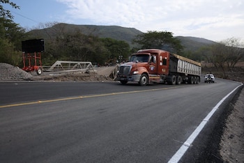 Un camión anaranjado con remolque avanza por una carretera asfaltada de dos carriles, cerca de cerros y vegetación