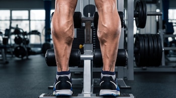 Piernas musculosas de una persona vista desde atrás, sobre una máquina de gimnasio. Se observan pantorrillas, venas marcadas, zapatillas deportivas y calcetines.