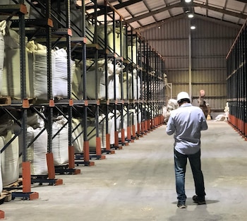 Un trabajador con casco de espaldas frente a estanterías metálicas altas cargadas con grandes sacos blancos dentro de una nave industrial