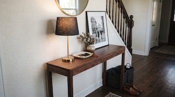 Una mesa consola de madera, lámpara con pantalla negra, espejo redondo, cuadro en blanco y negro, jarrón con ramas secas, cuenco con llaves, cesta y zapatos.