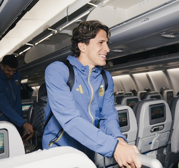 Un futbolista con chaqueta azul y amarilla de la selección argentina sonríe dentro de un avión, con otros asientos y un pasajero al fondo