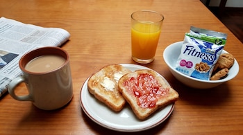 Vista cenital de una mesa de madera con un desayuno compuesto por café con leche, zumo de naranja, tostadas, cereales y galletas, junto a un periódico.
